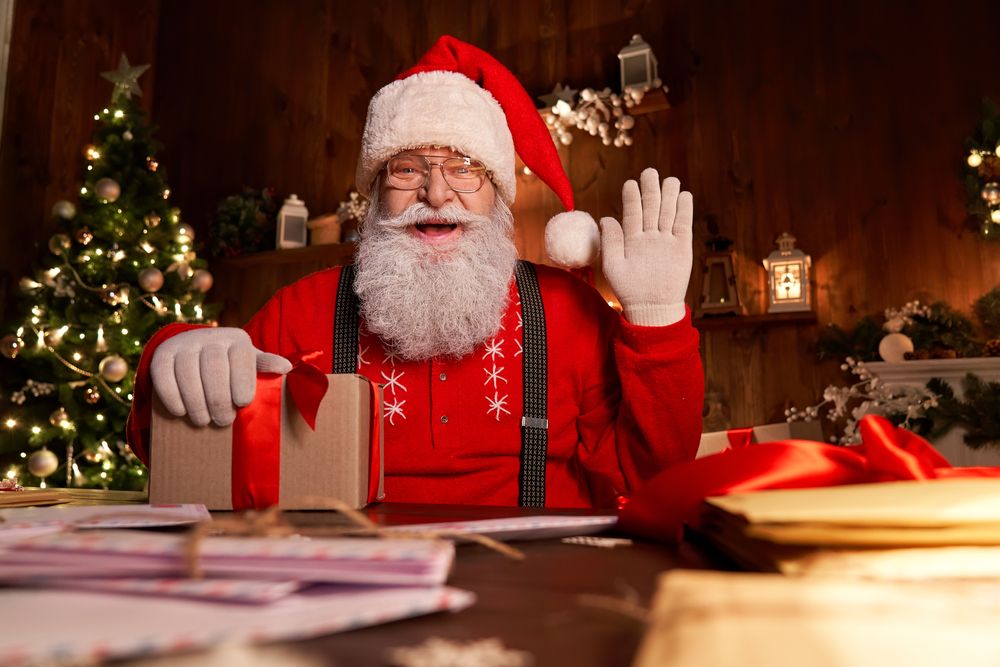Santa wrapping a present and waving in his workshop